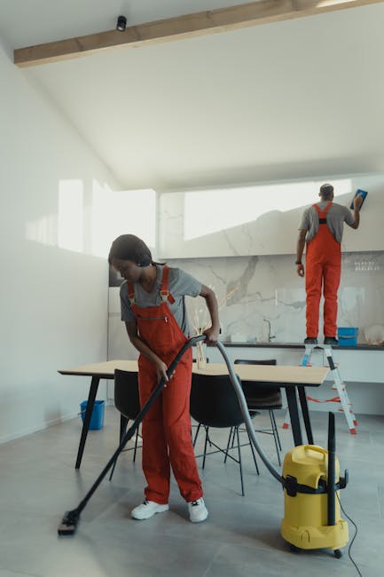 Two professional cleaners from Holborn Carpet Cleaners are conducting surface cleaning and maintenance in a modern kitchen with a minimalist design. The female cleaner is vacuuming the light wooden fl