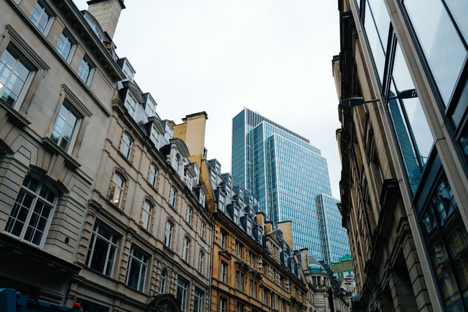 An urban street scene in Fleet Street, London, showing a mix of historic and modern buildings with brick and stone facades, large multi-pane windows, and decorative architectural details. A white stre