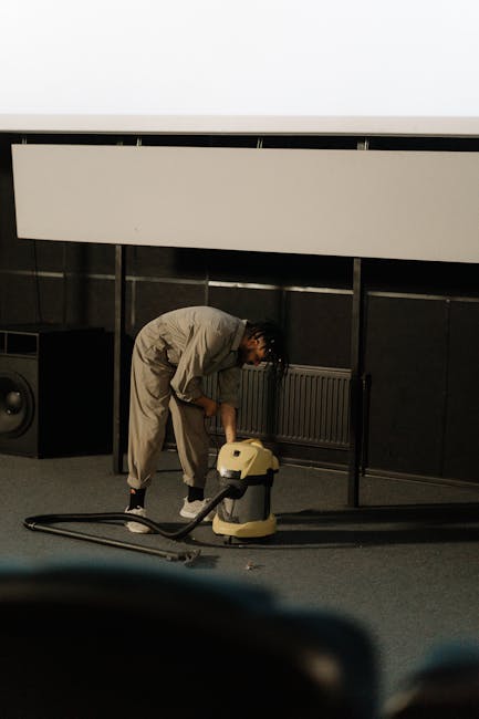 Close-up of a portable vacuum cleaner with a transparent dust container filled with debris, connected to a flexible, patterned hose attached to a black floor nozzle. The background shows a section of 